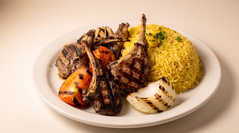 Grilled lamb chops, yellow rice, grilled tomatoes, and onion on a white plate.
