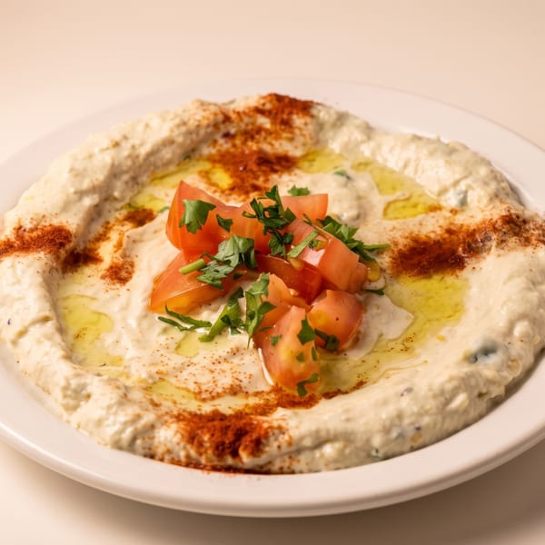 A white plate of hummus drizzled with olive oil, topped with diced tomatoes and parsley.