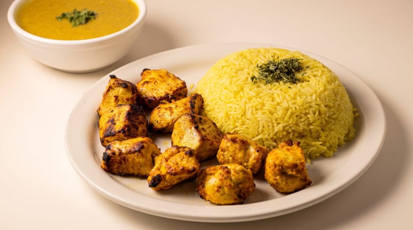 Plate of charred grilled chicken pieces, yellow rice, and a bowl of golden soup.